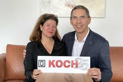 Christine Lieber-Koch und Michael Koch schauen freudig in die Kamera und halten das Schild 25 Jahre KOCH in den Händen.