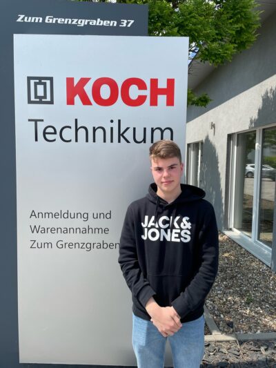 Finn Buschlinger steht vor dem Firmenschild der Michael Koch GmbH und lächelt freundlich in die Kamera.